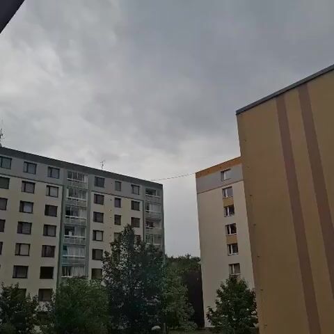 The video captures a cloudy sky above two apartment buildings, with trees visible in the foreground. One building is light beige with a vertical brown stripe, and the other is a light gray, tan, and green building. A thin, dark line, possibly a cable or wire, connects the two buildings at their upper levels. There are no people or any discernible activity.