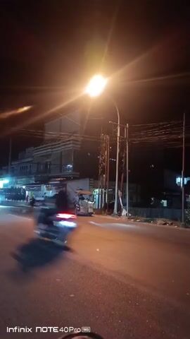 The video captures a nighttime street scene from the perspective of someone riding a motorcycle. A bright street lamp illuminates the asphalt road, casting long shadows. On the right side of the road, there are utility poles and wires. Across the street, a multi-story building with a striped facade is visible, with shops or businesses at the ground level. Several vehicles are present. A tuk-tuk is parked on the side of the road. Motorcycles are seen in motion, with headlights and taillights creating streaks of light. One motorcycle passes by, its rider partially visible. The overall atmosphere is one of a busy street at night, with traffic and illuminated businesses. The camera captures a reflection in what appears to be a motorcycle's rearview mirror at the bottom of the frame. The video has a timestamp overlay indicating it was recorded on an Infinix NOTE 40 Pro 5G device.