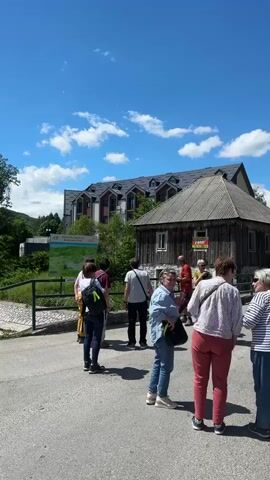 The video shows a group of people gathered outdoors on a paved area. In the background, there is a rustic wooden building with a weathered roof, and a more modern, multi-story building with a dark, angled roof. A large evergreen tree is prominent on the right side of the frame. Some of the people appear to be in conversation, while others are standing and looking around. The sky is blue with scattered white clouds, and the overall setting seems to be a daytime outdoor gathering.