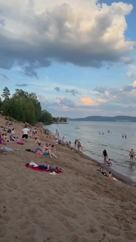 This is a vertical, medium shot of a crowded beach scene on a sunny day. The foreground is dominated by a sandy slope leading down to a calm body of water. Numerous people are scattered across the beach, some lying on towels, others sitting or walking. More people are in the water, swimming, wading, or standing near the shore. A line of trees forms a green backdrop on the left side of the frame, with hills visible in the distance on the right. The sky is a mix of blue and white clouds, with hints of pink and orange suggesting either sunrise or sunset. The water appears clear and shallow near the edge. The overall atmosphere is one of leisure and recreation.