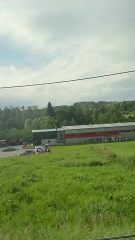 A POV video captures a landscape with a cloudy sky. The scene includes a building with red and gray elements and several vehicles in its vicinity. Further along, a white house with a dark roof is visible, bordered by a green hedge and a wire fence. A red sign with white text and an arrow is also present. Tall, dark green trees form a line on the right side of the frame.