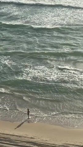 An aerial shot shows a person walking along a sandy beach next to a body of water with waves. The person is walking from left to right and appears to be heading towards the right side of the frame. The water is a greenish-blue color and the waves are breaking on the shore. The sky is not visible in the frame.