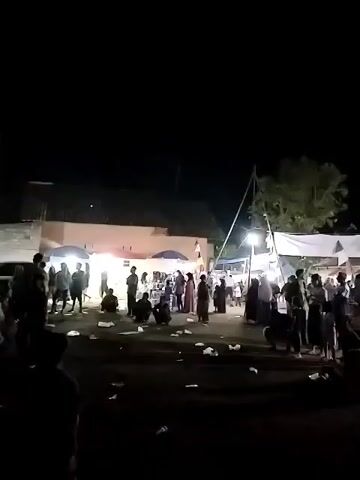 The video captures a nighttime scene at an outdoor gathering, possibly a festival or market. A crowd of people are visible, many of whom are silhouetted against the dimly lit background. Several tents or stalls are set up, adorned with colorful fabric draping. The ground appears to be dirt or unpaved, with scattered debris, possibly litter. Lights are illuminating some areas, creating a festive atmosphere. The sky is dark, indicating it is nighttime. There are trees in the background, partially obscured by the tents and darkness. The camera movement is slightly shaky, suggesting it is being held by hand.