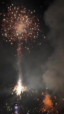 This video shows a nighttime fireworks display. The sky is dark, filled with smoke from the explosions. Bright bursts of light, predominantly in shades of yellow and white, erupt in the sky. Some fireworks leave trails of sparks as they ascend and descend. Below the fireworks, a crowd of people is visible, many holding up their phones to record the event. The scene appears to be outdoors, with some trees and structures faintly discernible in the background.