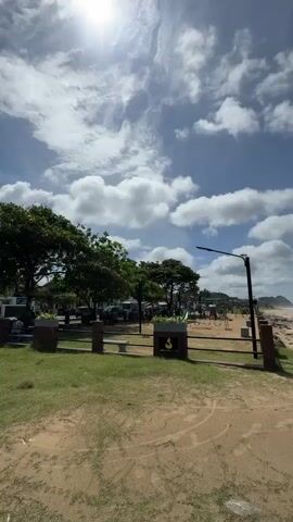 This is a vertical video of a sandy beach with a bright blue sky and scattered white clouds. In the foreground, there is a patch of green grass and a sandy path. A fence with brown posts and a green railing separates the grassy area from a playground with red equipment. Beyond the playground, there are trees and parked cars. The beach itself is lined with rocks and the ocean waves are visible. A street light is also present near the playground. The sun is shining brightly, casting a slight glare on the video. There are no people visible in the video.