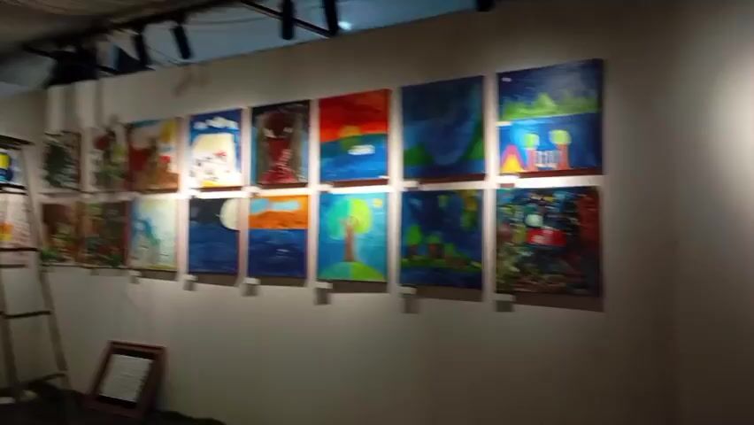 The video pans across an art exhibition. On the left wall, there are rows of paintings, mostly landscape and abstract art, with some featuring figures. An easel stands in the middle of the room, displaying a painting. On the right wall, more paintings are displayed in a grid pattern, showcasing a variety of colorful and childlike artworks. Hanging from the ceiling are decorative lanterns. The room has a white wall and a draped ceiling. There are a few chairs placed around the exhibition space. The lighting is provided by spotlights, casting shadows on the walls. Some books are scattered on the floor in the foreground. The overall impression is of a vibrant and child-friendly art display.