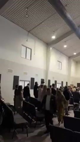 The video shows a large crowd of people gathered in a spacious, well-lit hall, which appears to be a church or auditorium. The people are standing, many are clapping, and some appear to be singing. At the front of the hall, a stage is visible with a large screen displaying information. At least two video cameras are positioned to record the event. Two men wearing 'MEDIA' shirts stand in front of them, operating the equipment. The ceiling is high and has a ribbed design, with industrial-looking ductwork running across it.