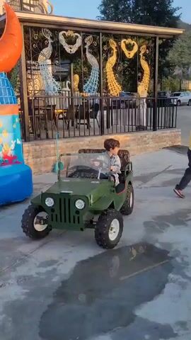 A young boy is driving a small green toy jeep. He is sitting in the driver's seat and holding the steering wheel. The jeep is moving forward on a paved surface. In the background, there is a building with decorative metalwork featuring peacock designs. There is also an inflatable structure to the left of the frame. The scene appears to be outdoors during daylight.