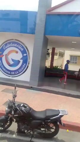 The video pans across a building with the logo "Gobernación Bolivariana de la Guaira" displayed on a white facade under a blue awning, where a person sits waiting. It then focuses on a corner with gray concrete structures and glimpses of a hilly landscape in the background. The view moves to a street corner with a yellow curb, a white building labeled "CORPOELEC," and two individuals unloading crates from a white truck. Finally, the video shows a tree and a post, again with the white building and hilly background visible.