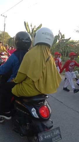 The video shows a group of people standing and riding motorcycles outdoors. In the background, several individuals in red and white uniforms are walking. In the foreground, a line of motorcycles is visible, with several people riding them. The scene appears to be a road with trees and vegetation on the side. The sky is clear and bright. The overall impression is of an outdoor gathering or event.