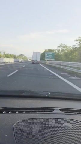 The video shows a view from inside a vehicle driving on a highway. The road is asphalt with white lane markings and a metal guardrail on the right side. A large green highway sign is visible on the right, indicating directions to Pescara, Teramo, Giulianova, and Mosciano S.A. A white truck is driving in front of the vehicle in the right lane. Further ahead, another white truck is visible. The sky is clear with some clouds. Trees and vegetation line the right side of the highway. The dashboard of the vehicle is visible in the foreground.