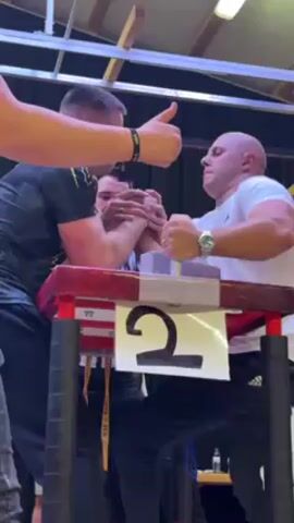 The video shows two men arm wrestling. One man has dark hair and a dark printed shirt and is on the left. The other man has a shaved head and a white shirt and is on the right. There is a referee in a striped shirt standing between them. They are arm wrestling on a red table with a large number 2 on it. The environment appears to be some sort of competition hall with a high ceiling. The man in the white shirt is struggling to win.