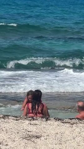 The video shows a beach scene with three people in shallow ocean water. In the foreground, there is a sandy beach with some seaweed. To the left, a young woman with blonde hair and a pink bikini top is partially submerged in the water, looking to her right. In the center, a woman with long dark hair wearing a red bikini top is also in the water, facing away from the camera. She appears to be interacting with the water, possibly splashing or gesturing. To the right, a man with a shaved head and wearing a dark shirt is visible from the shoulders up, facing the ocean. The ocean is blue and turquoise, with visible waves breaking closer to the shore. The sun appears to be bright, casting shadows.