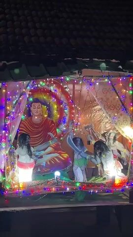 The video shows a Buddhist shrine under a dark roof. A colorfully lit cutout of the Buddha is in the center, with additional figures to his sides, also cutout and lit. There is a musical whirring noise. Colorful string lights frame the display.