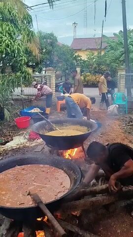 A group of men are cooking outdoors. In the foreground, two large woks are cooking over open fires. The wok on the left contains a dark, reddish-brown stew, and the wok on the right contains a lighter, yellowish stew. A man in a black shirt and shorts is kneeling by the wok on the right, tending to the fire. Another man in a yellow shirt and blue jeans is stirring the stew in the wok on the right with a long paddle. Behind them, several other men are gathered, with some preparing ingredients in blue and red buckets. The scene appears to be in a backyard or outdoor area, with trees and a fence visible in the background. A road and houses are also visible further in the background.