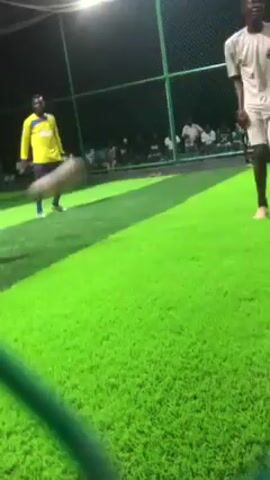 In an indoor soccer field, a person in a yellow shirt kicks a soccer ball. The soccer field has artificial green grass, and is surrounded by a green fence. There are spectators sitting along the fence. The indoor soccer field is lit by lights hanging from the ceiling. A person wearing a white shirt with the number 9 on the back is in the foreground, facing away from the camera.