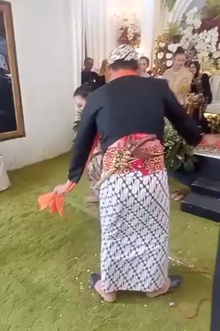 A wedding celebration takes place in a room with green carpeting and white drapes. In the foreground, a couple dressed in traditional Indonesian wedding attire dances. The woman wears a shimmering, long-sleeved top and a striped sarong, her hair styled in an updo. The man wears a black long-sleeved shirt, a sarong with a geometric pattern, and a patterned headcloth. An orange sash is draped over his left shoulder, and he holds a corner of it as they dance. A traditional dagger, a kris, is tucked into his waistband. In the background, several guests are seated and standing, observing the dance. There are floral decorations and white curtains adorning the area.