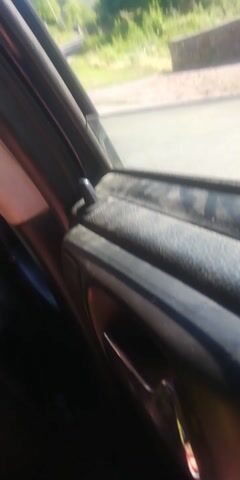 The video, shot from inside a vehicle, shows a view out of the driver's side window as the car travels along a road. The road curves to the right, bordered by a grassy embankment and trees on the left. A blue sign with text is visible on the embankment. The sky is clear and blue. The camera angle is tilted, making the scene appear sideways. The interior of the car, including a speaker and window trim, is visible in the foreground. The lighting suggests it is daytime.