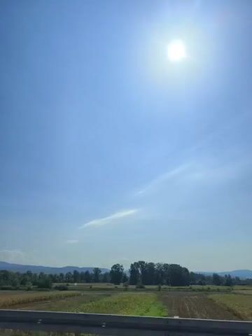 The video shows a bright, sunny day with a clear blue sky and a single, bright sun in the upper right quadrant. Below the sky, there is a landscape with fields, trees, and hills in the distance. In the foreground, a metal guardrail is visible, suggesting the viewpoint is from a moving vehicle on a road. The fields appear to be agricultural, with some areas showing harvested crops. The overall impression is of a rural, open environment under a clear sky.