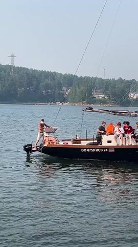 A man is standing at the back of a boat, holding a fishing net. He is wearing shorts and a hat. There are several other people on the boat, sitting down. The boat is on a body of water, and there is a forested shoreline in the background. The sky is hazy. The boat has the registration BO 0750 RUS 24.