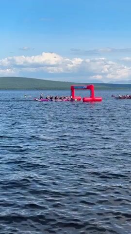 The video shows a wide body of water under a bright blue sky with scattered clouds. In the middle ground, there is a large inflatable red archway set up in the water, possibly as a starting or finishing line for a water event. Several people are visible on what appear to be paddleboards or small boats gathered around the inflatable arch. To the left, a lone person on a paddleboard is further out in the water, and to the right, another group of people on small boats or rafts are further away. The shoreline in the background consists of a tree-covered hill or mountain range. The water is choppy with visible ripples and waves.