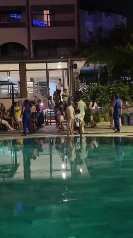 A group of people are gathered by a swimming pool at night. The scene takes place in Marmaris, Turkey, outside a building that appears to be a hotel or resort. The pool is illuminated with green lights, and the reflections of the people and the building are visible on the water's surface.

There are many people of various ages, some dressed in casual attire, others in swimwear. Some are dancing or moving to music, with hands in the air, suggesting a lively and celebratory atmosphere. Others are observing the activity, sitting or standing. The building behind them has several floors, with lights on in some of the windows. Neon blue lettering is visible on the upper floors, though the text is partially obscured and difficult to decipher. The presence of trees and foliage suggests an outdoor setting. The time of day is clearly night, with artificial lighting illuminating the area. The overall mood appears to be one of enjoyment and festivity.