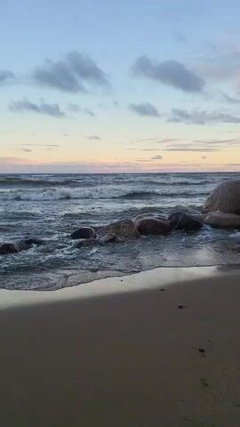 The video shows a beach scene with waves crashing against rocks and the shore. The sky is partly cloudy with a soft glow of sunset. The sand is damp from the receding tide. The waves are moderate in size and appear to be rolling in from a large body of water. There are no people visible in the video.