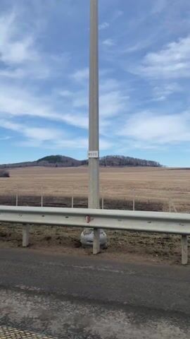 The video shows a view from a moving vehicle, likely a car, traveling along a paved road on a clear day. The sky is blue with scattered white clouds. To the right of the road is a guardrail. Beyond the guardrail, there is a large field of dry grass and vegetation. In the distance, there are two hills covered with sparse trees. The camera also captures road signs, including "no parking" and "video surveillance," attached to a metal pole.