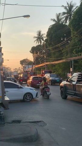 The video captures a street scene in Thailand at sunset, characterized by moderate traffic congestion. Various vehicles, including cars, trucks, and motorcycles, are visible on the asphalt road. Palm trees and other lush greenery line the side of the road. Several people are on motorcycles, and one individual is walking along the sidewalk. A white car is prominently positioned in the foreground. The sky is a gradient of orange and yellow, indicating the sunset. Overhead, a network of tangled power lines adds to the urban atmosphere.