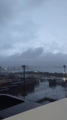 The video shows a rainy day view from a high vantage point overlooking a port. In the foreground, there is a white railing and part of a roof structure. Beyond that, there is a large open area with rows of stacked shipping containers and buildings. The ground appears wet from the rain, and there are some parked vehicles. In the middle ground, there is a body of water, likely a harbor or bay, with a distant cityscape visible across it. The sky is overcast and cloudy, indicating gloomy weather. Several tall light poles are also visible throughout the scene.