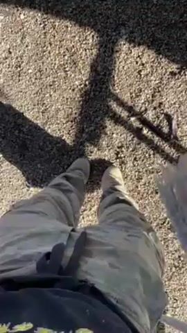 The video shows a first-person perspective of someone sitting on a swing outside. The person is wearing camouflage pants and tan-colored boots. The ground is covered in gravel and dirt. There are some construction materials visible in the background, including orange cones and wooden pallets. The camera moves slightly as the person swings.