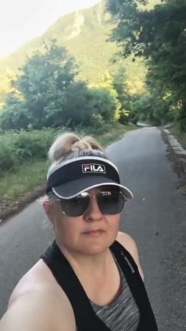This media file shows a woman with blonde hair in a bun, wearing a black Fila visor and sunglasses. She is also wearing a black tank top over a gray sports bra. She is outdoors on a paved path, likely in a wooded area, as trees and foliage are visible on either side. The lighting suggests it is daytime, possibly late afternoon or early evening, with dappled sunlight filtering through the trees. The woman appears to be walking or jogging, as the camera is moving with her. In the background, a black and white spotted dog on a leash is briefly visible, suggesting the woman is out for a walk or run with her pet. The overall mood seems relaxed and active, indicative of an outdoor recreational activity. The scene appears to be a natural, possibly rural setting. Visible text includes the brand name "FILA" on the visor and "Reebok" on the sports bra strap.