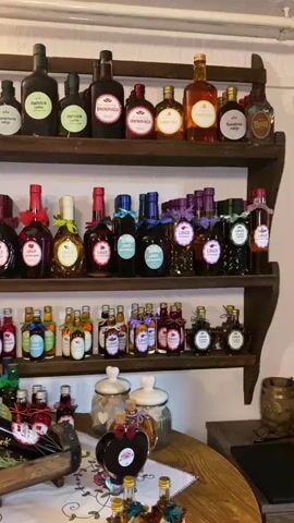 The video captures an interior scene, likely a shop or a tasting room, filled with numerous bottles of various spirits or liqueurs displayed on wooden shelves and a wooden table. The camera slowly pans from right to left, showcasing the arrangement.

On the top two wooden shelves, a wide assortment of bottles is neatly organized. The bottles vary in size, shape, and liquid color, ranging from clear to amber and dark red. Many bottles have distinctive labels with text. Visible text on the labels includes "MELVICA," "DRNOVAČA," "LINČURA," "KOŠĆELA RAJIJA," "BOROVICA," "PLANINSKA RAJIJA," "MEDENA RAJIJA," "ARONIJA," and "KADULJA," indicating different types of local spirits or herbal liqueurs. Some bottles are adorned with ribbons or decorative caps.

Below the shelves, a wooden table holds more bottles, some arranged in a wooden crate and a small barrel. There are also several decorative jars with cork stoppers, vases with yellow and pink flowers, and a small, heart-shaped bottle.

The room has a rustic aesthetic, with white textured walls and warm lighting from an unseen source and a visible lamp on the left. Decorative elements include a hanging antique-style scale with artificial fruit (pomegranates, grapes, apples), a framed certificate, and an old clock on a shelf to the left.

No people are explicitly visible, but conversations in a language that sounds like Croatian/Serbian/Bosnian are heard in the background throughout the video, suggesting visitors or staff are present off-camera. The overall ambiance indicates a place dedicated to showcasing and selling artisanal beverages.
