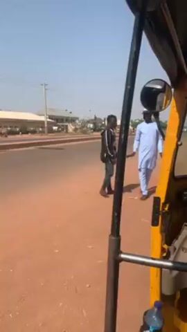 The video is shot from inside a three-wheeled vehicle, possibly a tuk-tuk or auto-rickshaw, parked on a red dirt road. Two men are standing on the road outside the vehicle, engaged in what appears to be a discussion. One man is wearing a black jacket with white patterns and dark pants. The other man is wearing a light blue traditional outfit. The driver or passenger inside the vehicle is wearing a red top and a beige knitted hat. The background shows a clear, blue sky and a building across the road. The camera slightly shifts, focusing on the driver/passenger as the vehicle starts moving. The initial focus on the two men outside blurs as they are left behind.