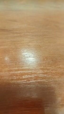 This is a close-up video of a wooden surface, likely a table. The wood grain is visible and appears to be a light to medium brown color. There are reflections on the surface, suggesting it is polished or varnished. The camera is held at a low angle, and the focus shifts slightly throughout the video, creating a shallow depth of field. The lighting appears to be coming from above and to the left, casting a bright highlight in the center of the frame. No people or other objects are visible in the video. The video is short, lasting only a few seconds.