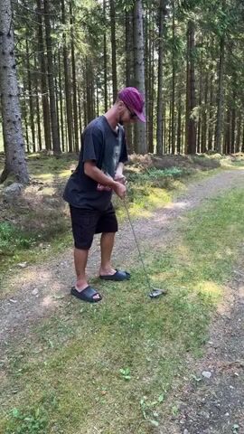 A man is playing golf in a forest clearing during the daytime. He is wearing a purple baseball cap, a dark t-shirt with a graphic print that includes text (partially visible as "Les lize Ranch"), black shorts, and black slide sandals. He holds a golf club and takes a swing at a small object on the grassy ground. The background is filled with tall trees, suggesting a dense forest, and there's a dirt path visible extending into the distance. The lighting indicates a sunny day.