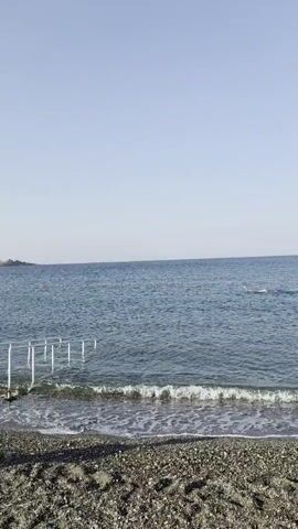 A video shows a beach with dark pebbles and a few white posts in the water. The sea is blue with white waves, and the sky is clear and light blue. In the distance, there are some buildings on a coastline.