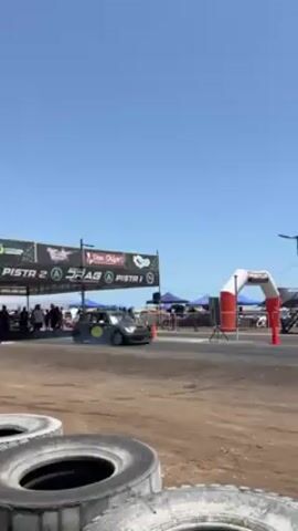A black Mini Cooper with a yellow logo on the door speeds down a drag strip under a clear blue sky. The car starts near a set of tires and a timing tower. A finish line archway can be seen further down the track. Tents line the track.