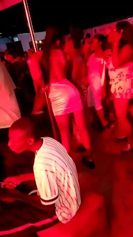 The video, taken from an eye-level perspective, shows a group of people dancing in a club or party setting illuminated by red lights. A man wearing a white striped baseball jersey is sitting on what appears to be a red cushion, working with something in his hands. He is positioned in the foreground and slightly to the left. Behind him, a crowd of people are dancing. A woman in the center wears a shiny silver dress. Another woman to her right is wearing a floral print outfit. A metal pole is visible on the left, and the overall atmosphere suggests a lively and festive occasion. The video is a little shaky.