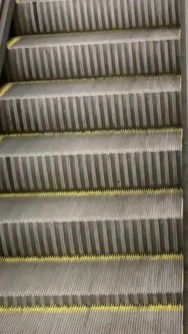 The video displays an upward-moving escalator from a low-angle perspective, focusing solely on the steps. No people are visible in the frame. The escalator steps are predominantly dark grey, featuring parallel, lighter grey ridged patterns that run horizontally across each step. A prominent bright yellow safety line marks the leading edge of each step, providing a visual cue for safe footing. The sides of the escalator, which would typically house the handrails and outer structure, appear dark, likely black, but the handrails themselves are not within the shot. The environment is indoors, characterized by consistent artificial lighting, suggesting a setting such as a shopping mall, subway station, or airport in L'hospitalet De Llobregat, Spain. The escalator is currently empty, moving continuously upwards without any passengers. There is no visible text in the video.