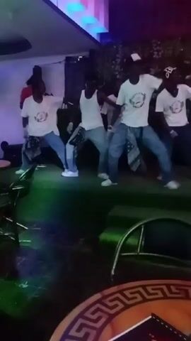 Here is a detailed description of the video:

The video captures a lively dance performance by a group of young people on an outdoor stage. Five individuals are prominently featured, all wearing similar outfits: white t-shirts with a graphic on the front, loose-fitting jeans, and white sneakers. One dancer sports a white baseball cap. 

The performance takes place on a stage covered with artificial grass. The stage is well-lit, and colorful LED lights add to the energetic atmosphere. The backdrop includes a white sheet or fabric. 

The foreground shows a glimpse of the venue's outdoor seating area, including a table with a patterned surface and chairs. Overall, the video conveys a scene of youthful energy, music, and dance in a festive environment.