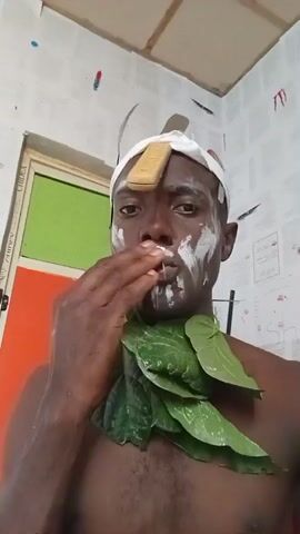 A man is shown from the chest up in a video that appears to be a selfie. He has dark skin and is shirtless. His face is partially covered with white substance, and he has a bandage wrapped around his head with a rectangular yellow object attached to it. He also has green leaves draped around his neck. He touches his face with his hand in the beginning, then salutes. The background consists of a white wall covered with papers, and a door with green and orange panels is visible on the left.