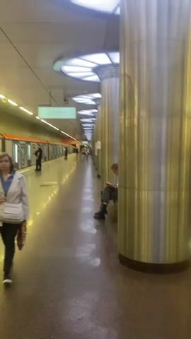 The media file captures a bustling scene on a subway platform in Lyubertsy, Russia. The camera's perspective is from the platform, looking down a long corridor lined with massive, fluted metallic columns. The ceiling is illuminated by bright, modern lighting, casting a warm glow on the polished floor, which reflects the lights and the movement of people and trains.

The primary subjects are the diverse group of people on the platform, some waiting for trains, others boarding or disembarking. In the foreground, a person is partially obscured by a large column, sitting down and seemingly waiting. Further down the platform, a train is visible, its doors open, with people entering and exiting. The train cars have a distinct orange stripe above the windows and doors, a common design element for public transport in some regions.

The activity taking place is the daily commute or travel via the subway system. The motion blur in some parts of the video, especially at the beginning, suggests movement, likely from the camera operator or a passing train, contributing to a dynamic and lively atmosphere. The lighting suggests it is likely daytime, or at least the station is well-lit, typical of underground or enclosed public spaces.

Notable details include the architectural style of the station with its large columns and modern lighting. The presence of multiple individuals, dressed in casual attire, indicates a public transportation hub. The reflections on the floor add a sense of depth and polish to the scene. The overall emotion conveyed is one of everyday activity and the hustle and bustle of urban life. No visible text is discernable in the provided frames.