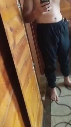 A man is standing in front of a mirror in a room with wooden walls and a bed. He is shirtless and wearing black pants. He is holding a phone in his left hand and flexing his right bicep. The camera is held by the man, taking a selfie.