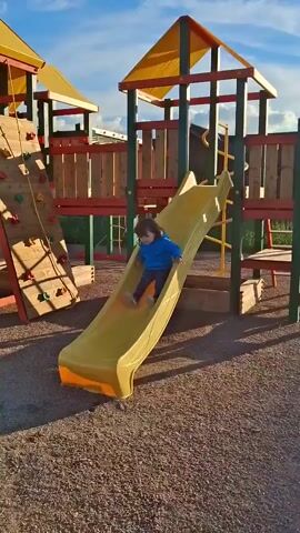 A child wearing a blue hoodie and patterned leggings slides down a yellow playground slide. They land on the ground and then run towards the camera, laughing and raising an arm. The playground features a climbing wall and other structures. The ground is covered in gravel. The sun is setting, casting long shadows.