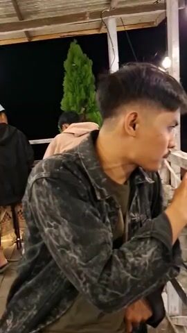 The video shows an outdoor seating area of a restaurant or cafe at night. The scene is illuminated by string lights and lamps. In the foreground, a young man is seen drinking from a glass of iced beverage, and there is a plate of sausages on skewers and a smartphone on a table. The camera pans to reveal the rest of the establishment, which has multiple levels with outdoor seating. People are seated at tables and on benches, some appear to be dining, others are socializing. A set of stairs leads to the upper level where more seating and tables are visible. The ground is covered in gravel, and there are some plants and trees around the area. Motorcycles are parked in the background.