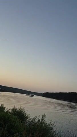 The video shows a wide river with a boat in the middle. The sky is clear and light blue, with a hint of pink at the horizon. Trees line the banks of the river. The boat appears to be a passenger boat, and it is moving at a moderate speed. The water is calm, with small ripples on the surface. The overall impression is one of peace and tranquility.