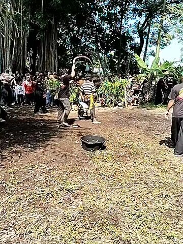 The video shows two men in a yard with a crowd of people surrounding them. The two men appear to be performing a dance or martial art form, one holding a whip that he swings around. One man is wearing a black shirt and dark pants, and the other is wearing a striped shirt and dark pants with a yellow belt. There is a black bucket on the ground near them.  The yard is covered in dry grass and is surrounded by trees and bushes. The crowd of people appears to be watching the performance.