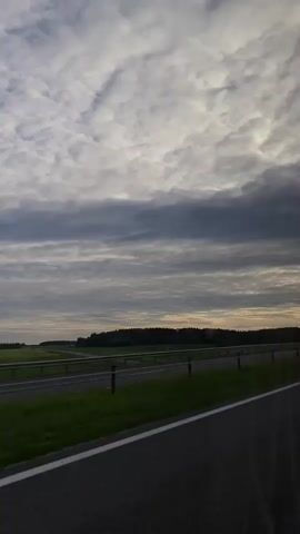 This is a vertical, low-angle, first-person perspective video filmed from a moving vehicle. The camera is looking forward and slightly to the left. The foreground is dominated by the dark asphalt of a highway lane, with a white line marking the edge of the lane. A metal guardrail separates the highway from a grassy median and a field beyond. In the distance, a line of trees forms a dark horizon under a cloudy sky. A white van towing a trailer is visible in the middle ground, traveling in the same direction as the camera vehicle. The sky is filled with a thick layer of textured, gray and white clouds, with hints of pale light breaking through in the distance. The overall mood is somewhat overcast and possibly early morning or late evening.