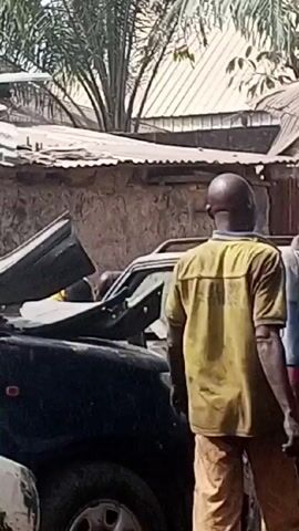 The video shows a man standing near two severely damaged cars in an outdoor environment. The man is Black with a shaved head, wearing a short-sleeved yellow shirt and tan pants. He is looking at the vehicles. One of the cars, a dark color, has extensive front-end damage. The other, painted blue, also appears heavily damaged. The background reveals a cluttered scene with what appears to be a building constructed from corrugated metal and various indistinct objects. The video also shows the underside and side of a white truck, which is heavily scratched and has a sign that says "odo City" on its side.